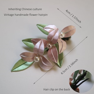 Puede incluir: Una horquilla de flores vintage hecha a mano. El accesorio para el cabello presenta flores de tela rosa con detalles de perlas y hojas verdes. La horquilla mide 6,5 cm de largo y 4 cm de ancho. El texto en la imagen dice: Inheriting Chinese culture, Vintage handmade flower hairpin.