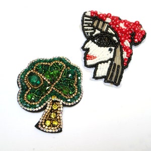 May include: Two beaded patches, one depicting a green shamrock-shaped tree and the other a woman's face with a red and white striped hat.