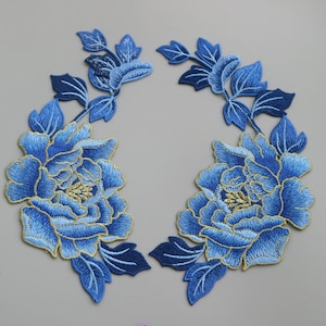 Puede incluir: Dos apliques florales bordados en azul con detalles dorados. Cada aplique presenta una flor grande con flores y hojas más pequeñas.
