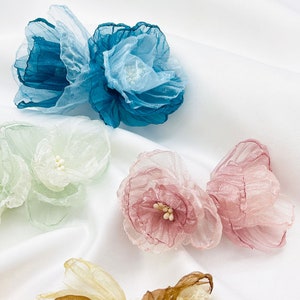May include: Assortment of four handmade fabric flowers in various colours, including teal, light green, pink, and beige. Each flower is made of layered tulle and has a delicate, ruffled texture.