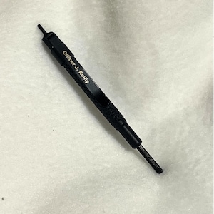 May include: A black multi-tool with the text "Officer J. Reilly" printed on the side. The tool has a textured grip and a pointed end. The tool is lying on a white, textured surface.