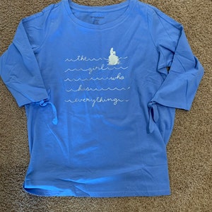 May include: A light blue long-sleeved t-shirt with a white graphic of a mermaid on a rock with waves and the text "the girl who has everything".