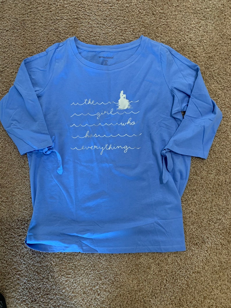 May include: A light blue long-sleeved t-shirt with a white graphic of a mermaid on a rock with waves and the text "the girl who has everything".