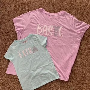 Puede incluir: Dos camisetas, una azul claro y otra rosa claro, con un dise&ntilde;o gr&aacute;fico en cada una. La camiseta azul claro dice "EXTRA" con una silueta del castillo de Cenicienta. La camiseta rosa claro dice "BASIC" con una silueta de la cabeza de Mickey Mouse y una taza.
