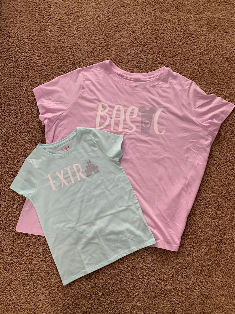 Puede incluir: Dos camisetas, una azul claro y otra rosa claro, con un dise&ntilde;o gr&aacute;fico en cada una. La camiseta azul claro dice "EXTRA" con una silueta del castillo de Cenicienta. La camiseta rosa claro dice "BASIC" con una silueta de la cabeza de Mickey Mouse y una taza.