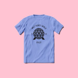Pu&ograve; includere: Una maglietta blu chiaro con una grafica nera del giro Spaceship Earth ad Epcot. La grafica include il testo "Walt Disney World 19 82 Epcot".
