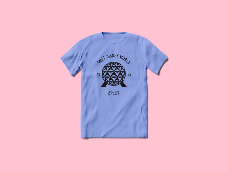 Pu&ograve; includere: Una maglietta blu chiaro con una grafica nera del giro Spaceship Earth ad Epcot. La grafica include il testo "Walt Disney World 19 82 Epcot".