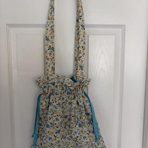 May include: A floral drawstring bag with a cream background and blue and yellow flowers. The bag has long straps and a light blue drawstring closure. The bag is hanging on a white door.