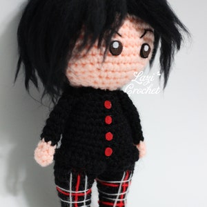 May include: Crocheted doll with black hair, a black and red plaid outfit, and red buttons. The doll is wearing a black jacket with red buttons and black trousers with a red and white plaid pattern. The doll is standing on a white surface. "Lazy Crochet" is written on the doll's chest.