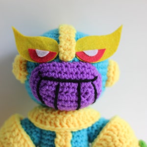 Puede incluir: Un juguete de peluche tejido a crochet de Thanos del universo Marvel. El juguete es azul, amarillo y morado con una boca negra y ojos rojos. El juguete tiene una diadema amarilla de fieltro con ojos rojos.