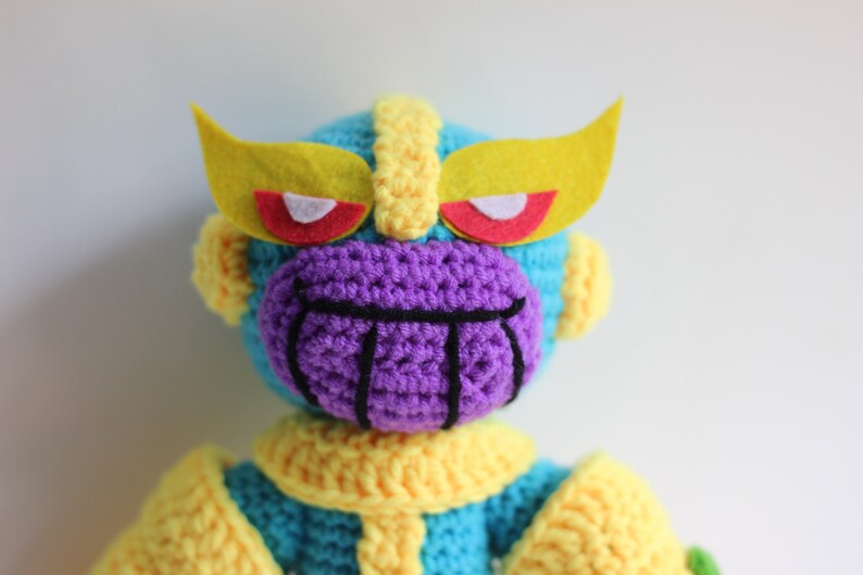 Puede incluir: Un juguete de peluche tejido a crochet de Thanos del universo Marvel. El juguete es azul, amarillo y morado con una boca negra y ojos rojos. El juguete tiene una diadema amarilla de fieltro con ojos rojos.