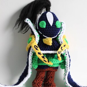Puede incluir: Un juguete de peluche tejido a crochet de un personaje con ojos verdes, una coleta negra y un atuendo amarillo, verde y azul. El juguete está de pie sobre una superficie blanca. El texto "Lazy Crochet" es visible en la parte inferior del juguete.
