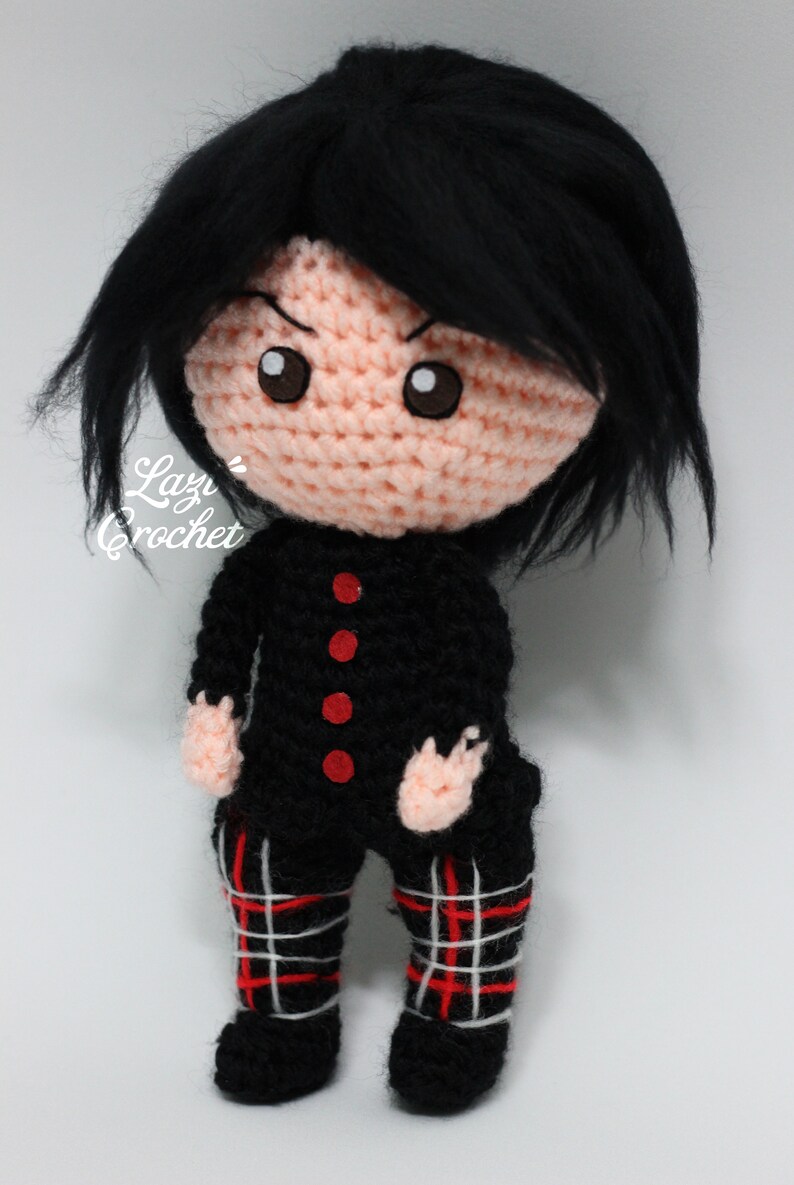 May include: A crocheted doll with black hair, a black and red tartan skirt, and a black shirt with red buttons. The doll has a pink face with brown eyes and a serious expression.
