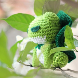 Op de afbeelding: Groen gehaakt amigurumi speelgoed met een wit gezicht en groene en blauwe bladeren op de achtergrond. Het speelgoed heeft de tekst "Lizzi Crochet" erop.