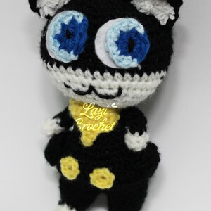 May include: A black and white crocheted plush toy cat with blue eyes and yellow accents. The toy is wearing a yellow scarf and has a white crocheted face.