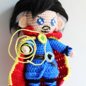 Könnte beinhalten: Ein gehäkeltes Plüschtier von Doctor Strange, einem Superhelden aus Marvel Comics. Das Spielzeug trägt ein blaues und rotes Kostüm mit einem gelben Umhang und hält einen gelben Faden in seiner rechten Hand. Das Spielzeug hat eine schwarze Perücke und blaue Augen.