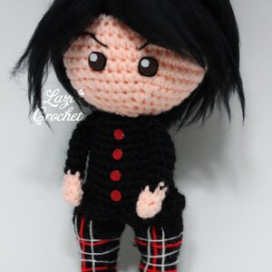 May include: A crocheted doll with black hair, a black and red tartan skirt, and a black shirt with red buttons. The doll has a pink face with brown eyes and a serious expression.