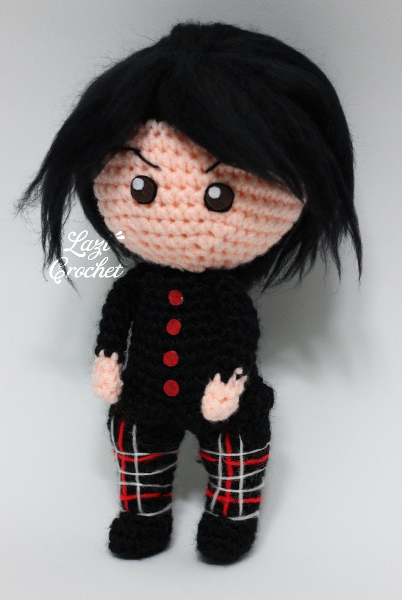 May include: A crocheted doll with black hair, a black and red tartan skirt, and a black shirt with red buttons. The doll has a pink face with brown eyes and a serious expression.