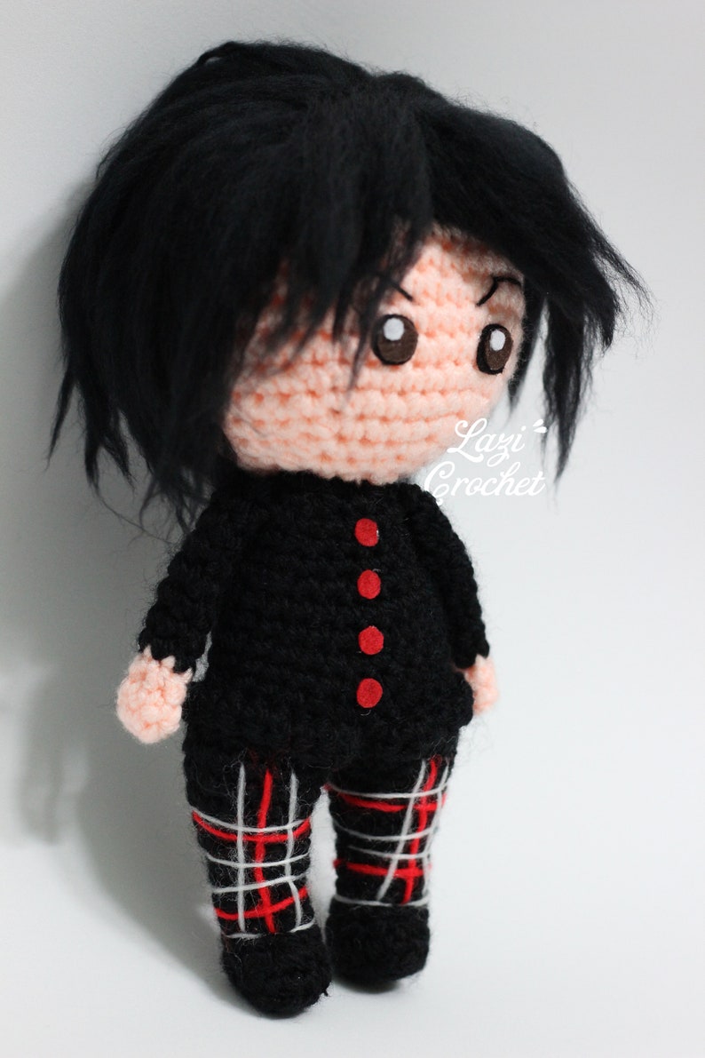 May include: Crocheted doll with black hair, a black sweater with red buttons, and red and white plaid trousers. The doll is standing on a white surface. The text "Lazy Crochet" is visible on the doll's right shoulder.