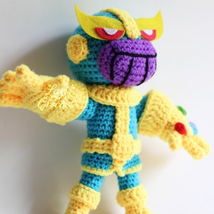 Puede incluir: Juguete de peluche Thanos tejido a crochet con una cara morada, un cuerpo amarillo y azul y un guantelete del infinito amarillo. El juguete est&aacute; de pie sobre un fondo blanco.