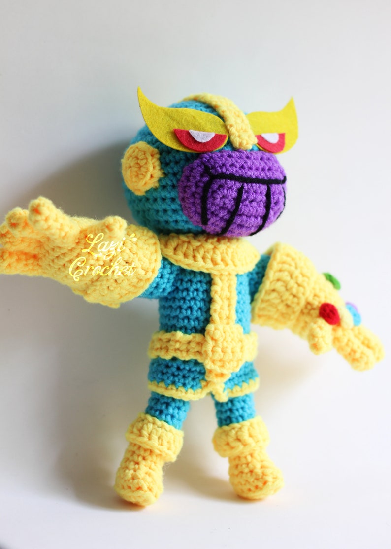 Puede incluir: Juguete de peluche Thanos tejido a crochet con una cara morada, un cuerpo amarillo y azul y un guantelete del infinito amarillo. El juguete est&aacute; de pie sobre un fondo blanco.