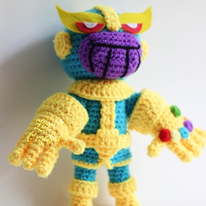 Puede incluir: Un juguete de Thanos tejido a crochet con una cara morada, guantes amarillos y un cuerpo azul. El juguete lleva un casco amarillo con ojos rojos y blancos. El juguete sostiene el Guantelete del Infinito en su mano derecha.