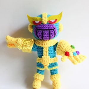 Puede incluir: Un juguete de peluche tejido a crochet de Thanos del Universo Cinematogr&aacute;fico de Marvel. El juguete es azul y amarillo con una cara morada y un Guantelete del Infinito dorado. El juguete lleva una diadema amarilla con ojos rojos.