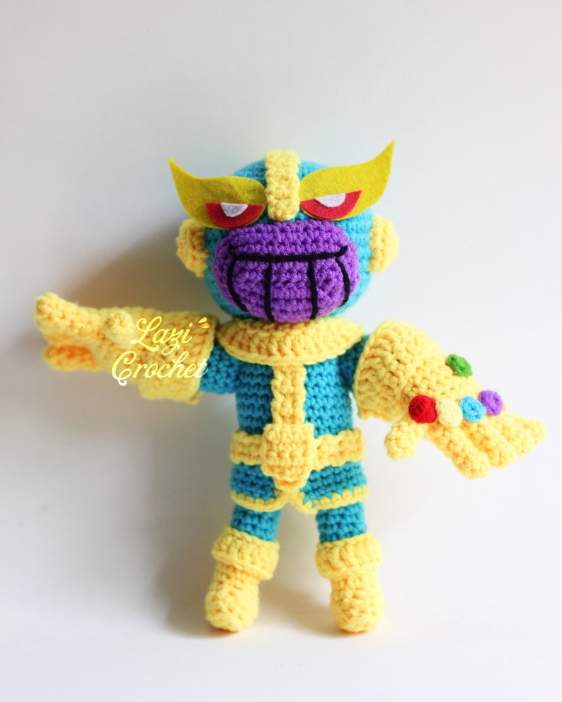 Puede incluir: Un juguete de peluche tejido a crochet de Thanos del Universo Cinematogr&aacute;fico de Marvel. El juguete es azul y amarillo con una cara morada y un Guantelete del Infinito dorado. El juguete lleva una diadema amarilla con ojos rojos.