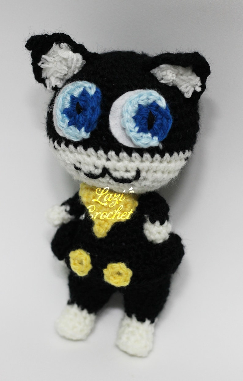 May include: A black and white crocheted plush toy cat with blue eyes and yellow accents. The toy is wearing a yellow scarf and has a white crocheted face.