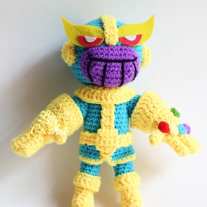 Könnte beinhalten: Ein gehäkeltes Plüschtier von Thanos aus dem Marvel-Universum. Das Spielzeug ist blau, gelb und lila mit einem lila Gesicht und gelben Augen. Das Spielzeug trägt einen gelben Infinity-Handschuh mit bunten Edelsteinen.