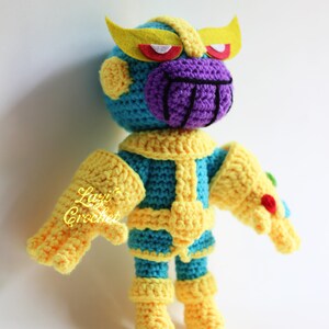 Puede incluir: Un juguete de Thanos tejido a crochet con una cara morada, ojos amarillos y un guantelete del infinito amarillo. El juguete lleva un traje azul y amarillo.