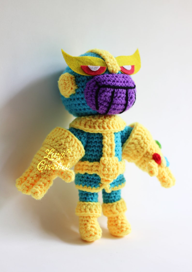 Puede incluir: Un juguete de Thanos tejido a crochet con una cara morada, ojos amarillos y un guantelete del infinito amarillo. El juguete lleva un traje azul y amarillo.
