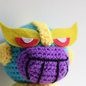Puede incluir: Juguete de peluche tejido a crochet con una cara morada, detalles amarillos y azules y ojos de fieltro rojo y blanco.