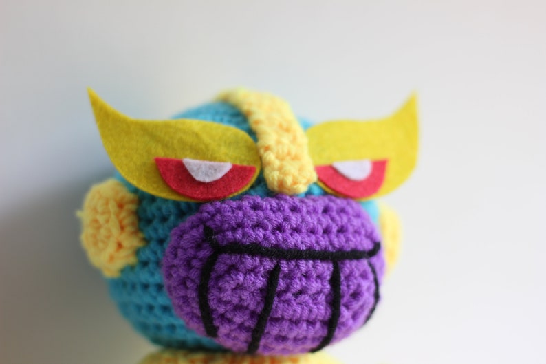 Puede incluir: Juguete de peluche tejido a crochet con una cara morada, detalles amarillos y azules y ojos de fieltro rojo y blanco.