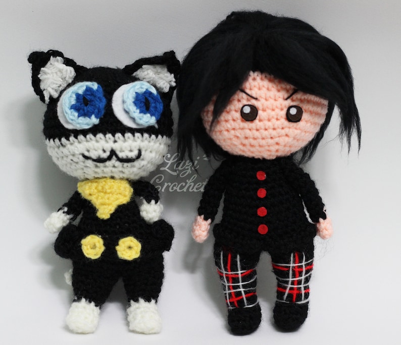 May include: Crocheted plush toys of a black cat with blue eyes and a human character with black hair and red buttons. The cat is wearing a yellow collar and black trousers. The human character is wearing a black shirt and red and white checked trousers.