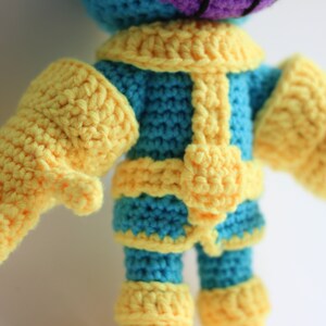 Puede incluir: Juguete de peluche tejido a crochet de un personaje de piel morada con guantes amarillos y un atuendo azul y amarillo.