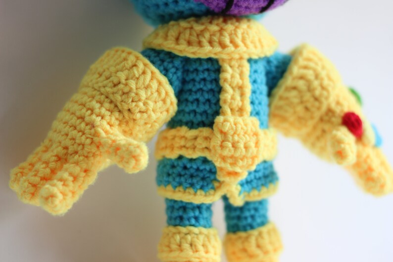 Puede incluir: Juguete de peluche tejido a crochet de un personaje de piel morada con guantes amarillos y un atuendo azul y amarillo.