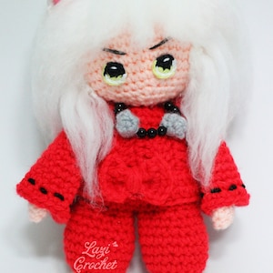 Puede incluir: Un juguete de peluche tejido a crochet de un personaje con cabello blanco, ropa roja y ojos negros. El juguete tiene una corbata de lazo y un collar de cuentas negras. El juguete está etiquetado como "Lazi Crochet".