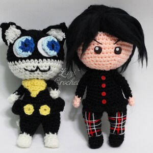 May include: Crocheted plush toys of a black cat with blue eyes and a human character with black hair and red buttons. The cat is wearing a yellow collar and black trousers. The human character is wearing a black shirt and red and white checked trousers.