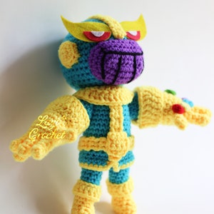 Puede incluir: Un juguete de peluche tejido a crochet de Thanos del Universo Cinematogr&aacute;fico de Marvel. El juguete lleva un traje amarillo y azul con una cara morada y cejas amarillas. El juguete sostiene un guante amarillo con gemas.