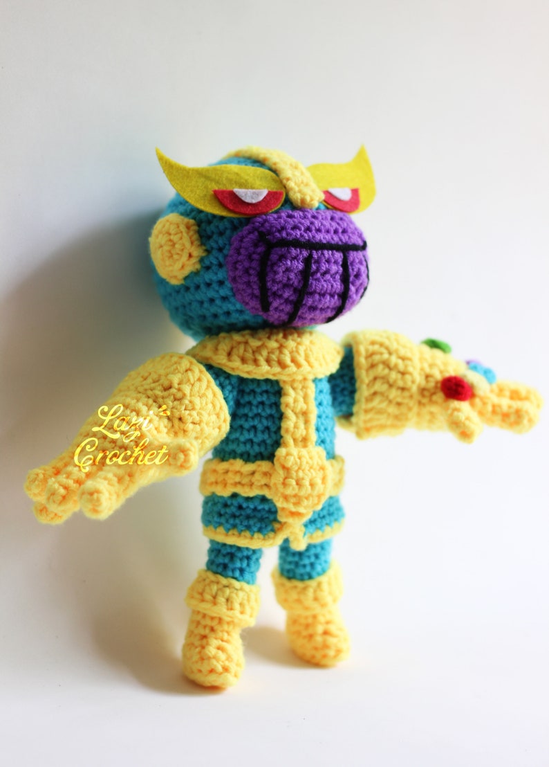 Puede incluir: Un juguete de peluche tejido a crochet de Thanos del Universo Cinematogr&aacute;fico de Marvel. El juguete lleva un traje amarillo y azul con una cara morada y cejas amarillas. El juguete sostiene un guante amarillo con gemas.