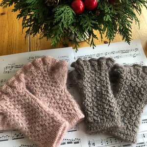 Könnte beinhalten: Zwei Paar gestrickte Fingerhandschuhe in zartem Rosa und Taupe mit einem strukturierten Muster. Die Handschuhe liegen auf Notenblättern, im Hintergrund eine festliche grün-rote Dekoration.