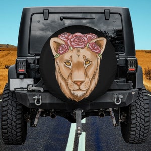 May include: A black spare tire cover with a brown lion's face wearing a pink flower crown. The lion has a pink nose and brown eyes.
