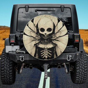 May include: A black Jeep with a spare tire cover featuring a skeletal figure with wings. The figure has a skull face with a toothy grin and is wearing a tattered dress. The background is a blurred image of a road and sky.