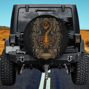 Puede incluir: Una cubierta de rueda de repuesto negra con un diseño de escorpión dorado y la palabra "Scorpion" en una pancarta. El escorpión está rodeado de detalles florales ornamentados y el símbolo astrológico de Escorpio.