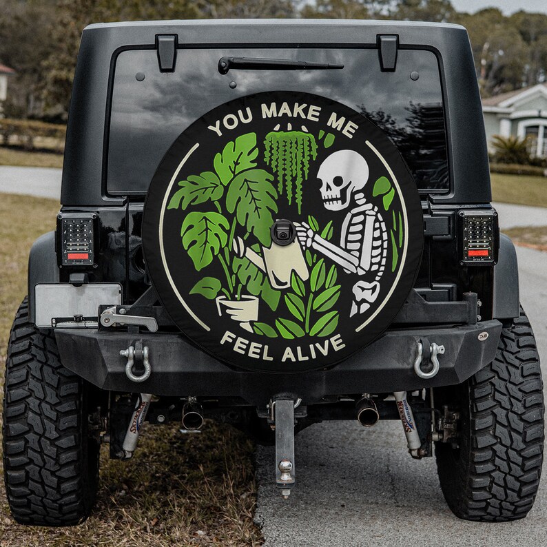 May include: A black Jeep with a spare tire cover that says "You Make Me Feel Alive". The cover features a skeleton watering a plant with a watering can.