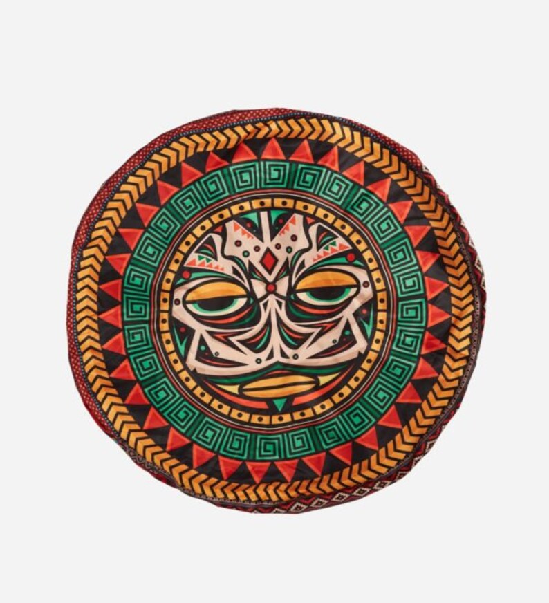 May include: A round, colorful cushion with a tribal design featuring a stylized face with geometric patterns. The cushion is primarily red, orange, green, and black.
