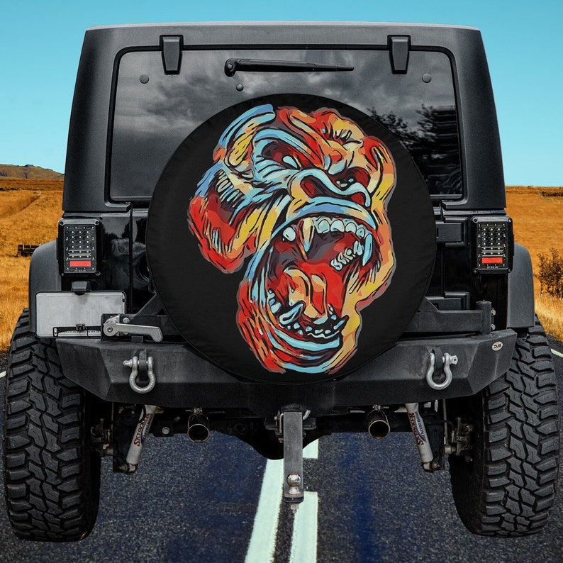 May include: A black spare tire cover with a colorful illustration of a gorilla's face with its mouth open, showing teeth. The gorilla's face is painted in shades of red, yellow, blue, and white.