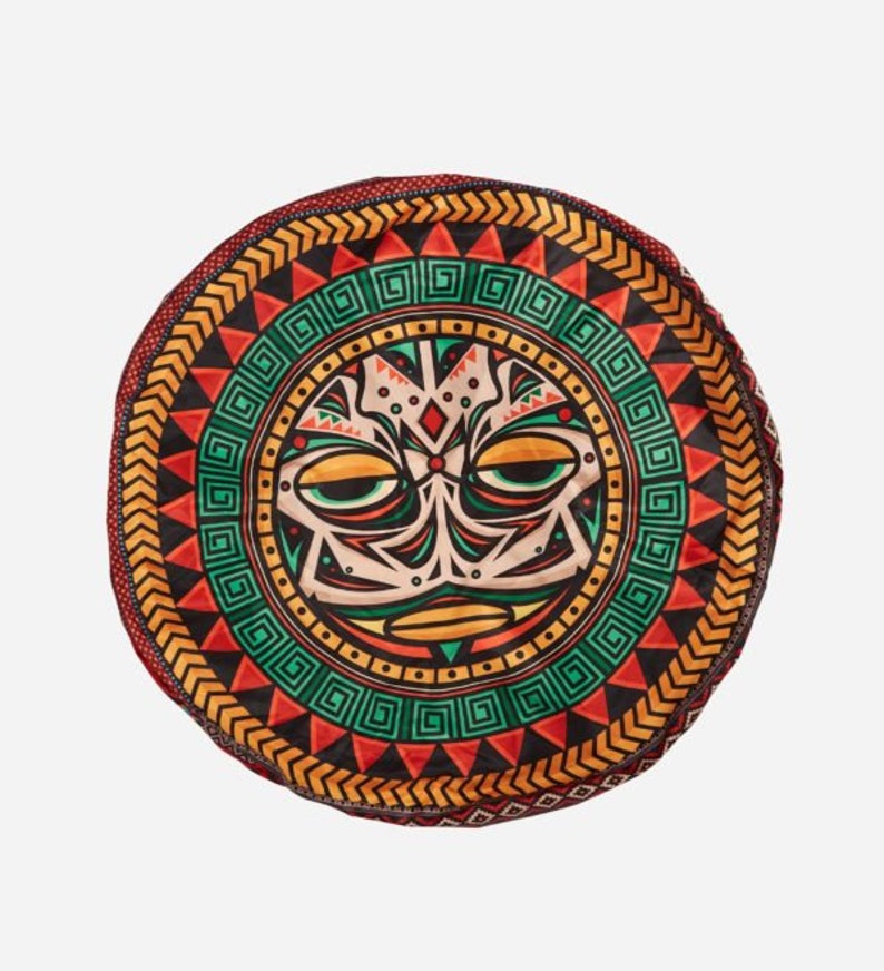 May include: A round, colorful cushion with a tribal design. The center of the cushion features a stylized face with geometric patterns in shades of red, green, yellow, and black.