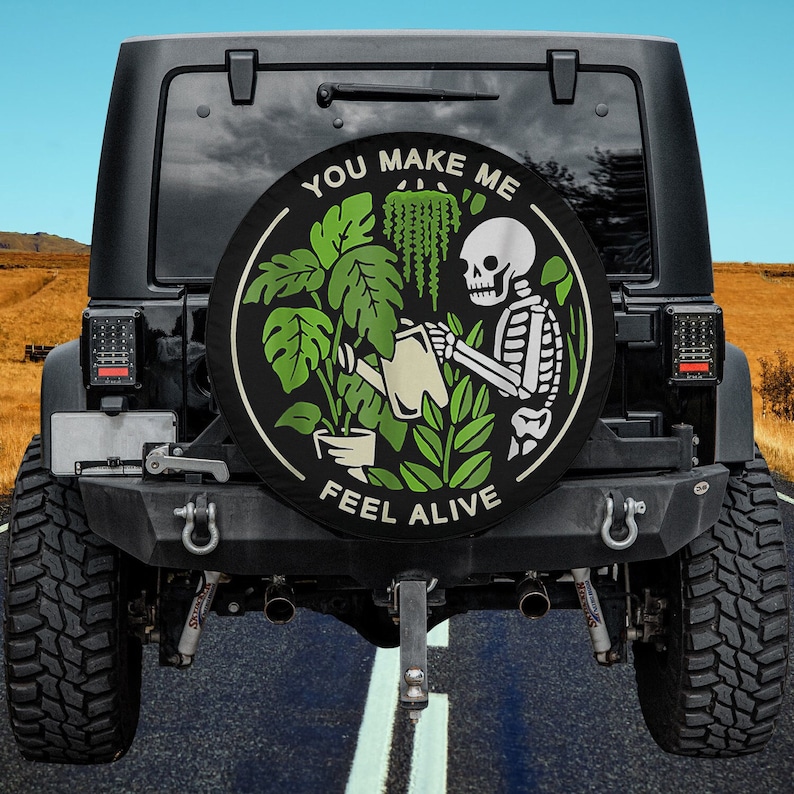 May include: A black spare tire cover with a white skeleton watering a green plant. The cover has the text "You Make Me Feel Alive".